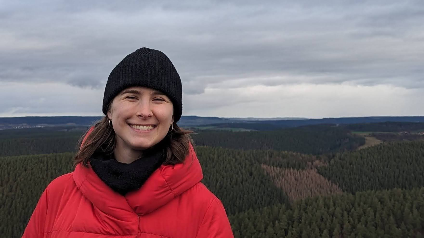 A photo of Rufio in a coat and a hat with an outlook over a forest behind her.